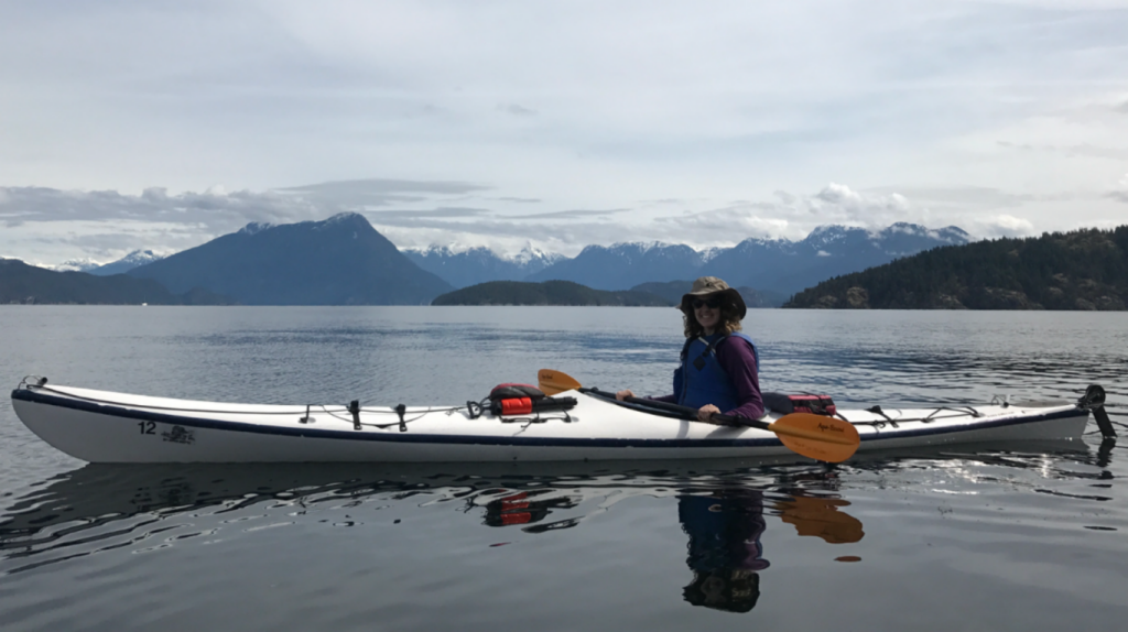 Kaitlyn smiling at the camera while sitting in a kayak that is in a large body of water. Mountains are in the distant background.